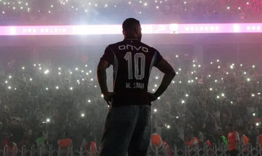 Back-view of a man in a black jersey with the number 10 on stage, hands on hips, facing a cheering crowd at a concert with many phone lights.