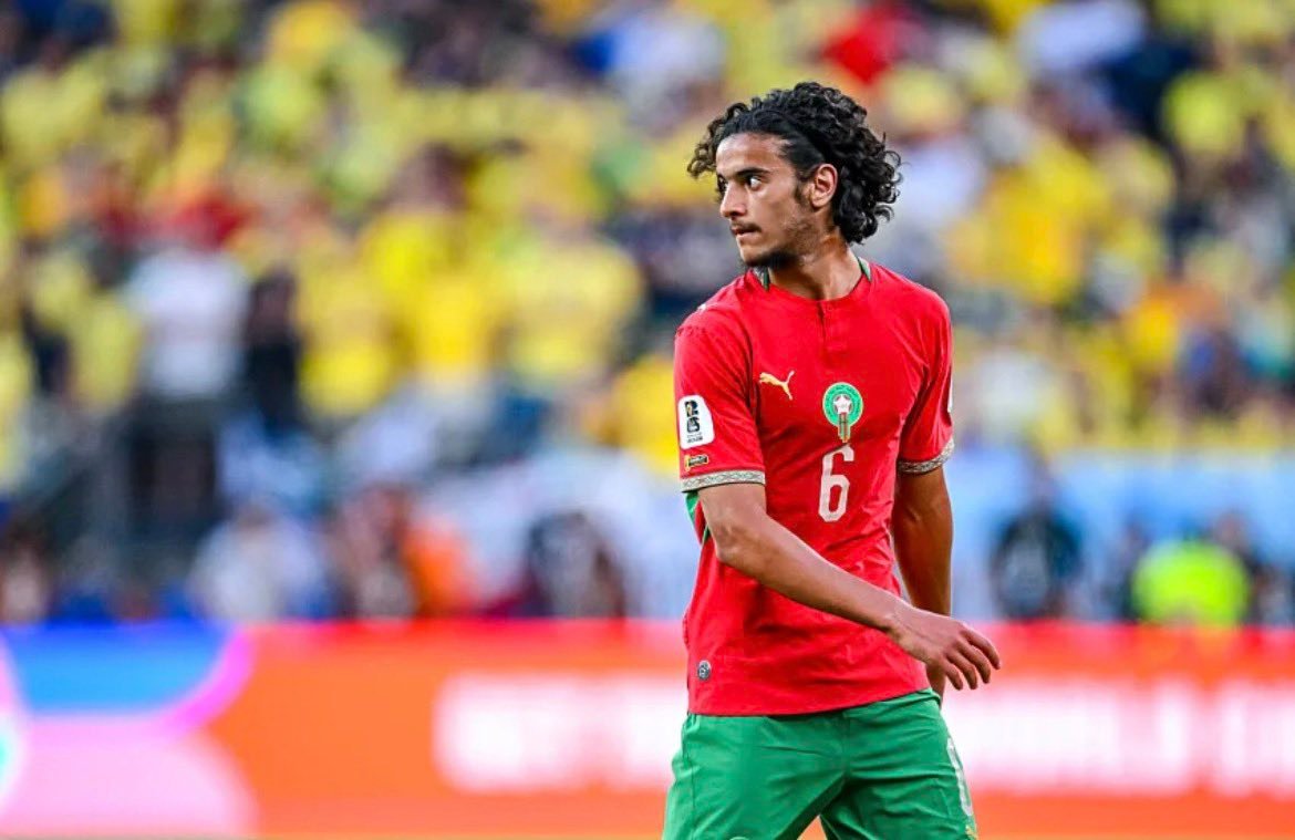 Soccer player in a red jersey with number 6, standing on the pitch with a yellow-clad crowd behind him.