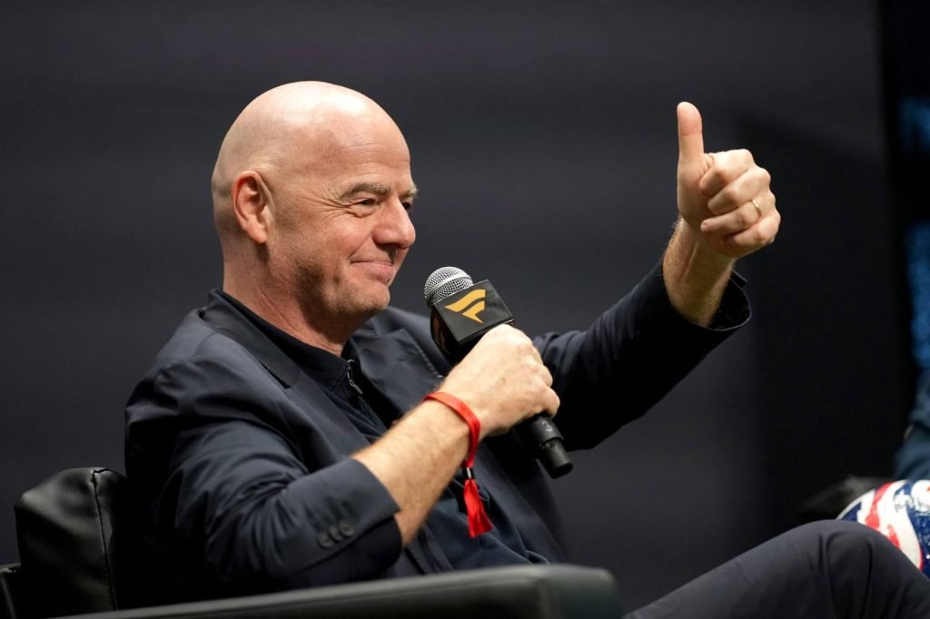 Bald man in a dark suit speaks into a microphone while giving a thumbs-up gesture on stage.