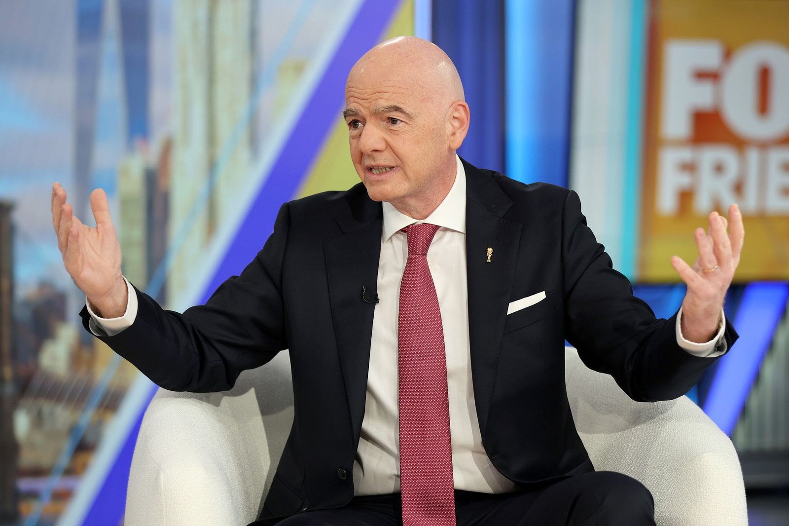 Bald man in a dark suit and red tie speaks while gesturing with his hands on a colorful TV set background.