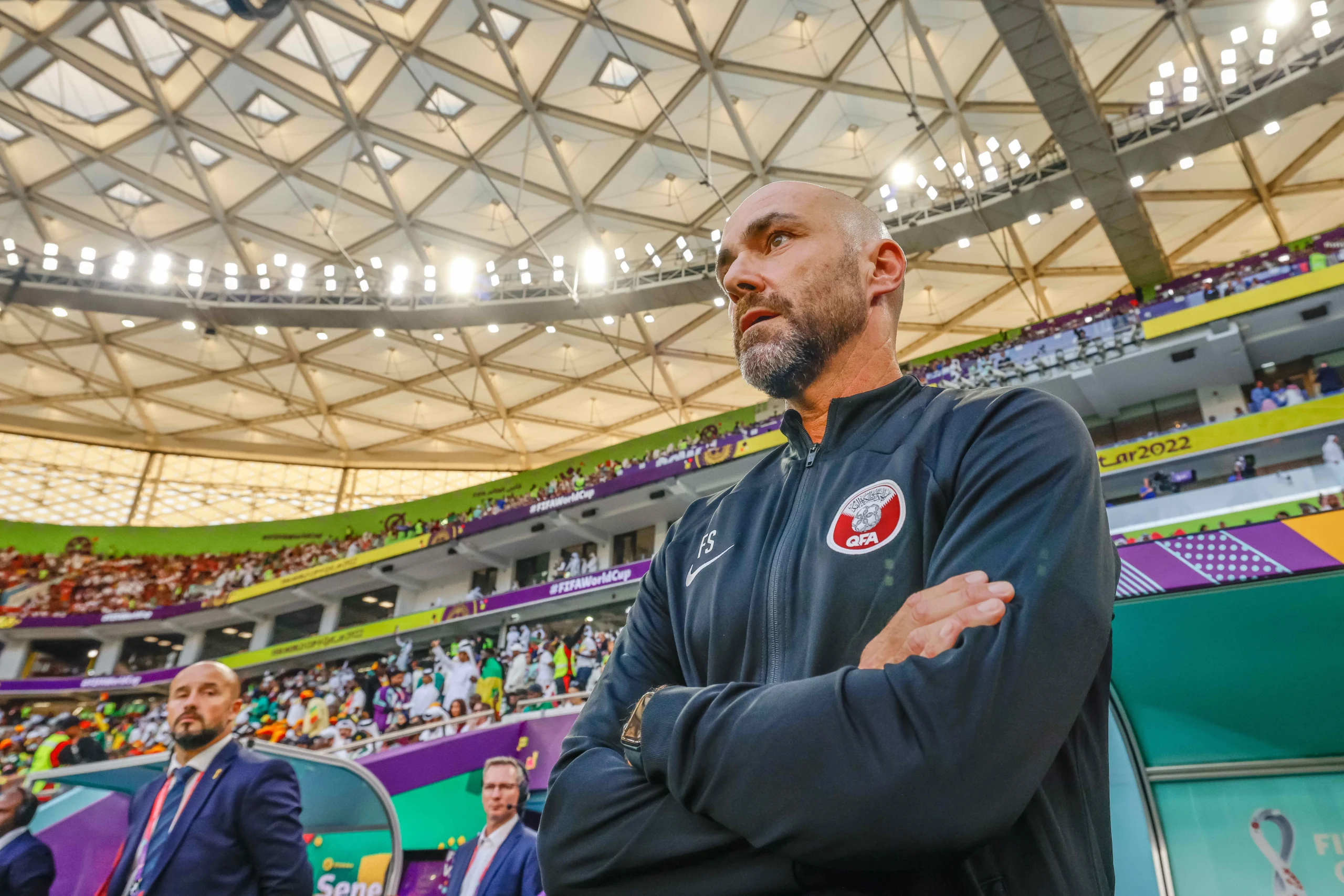Man in a dark jacket with a QFA badge stands courtside in a large indoor stadium with spectators.