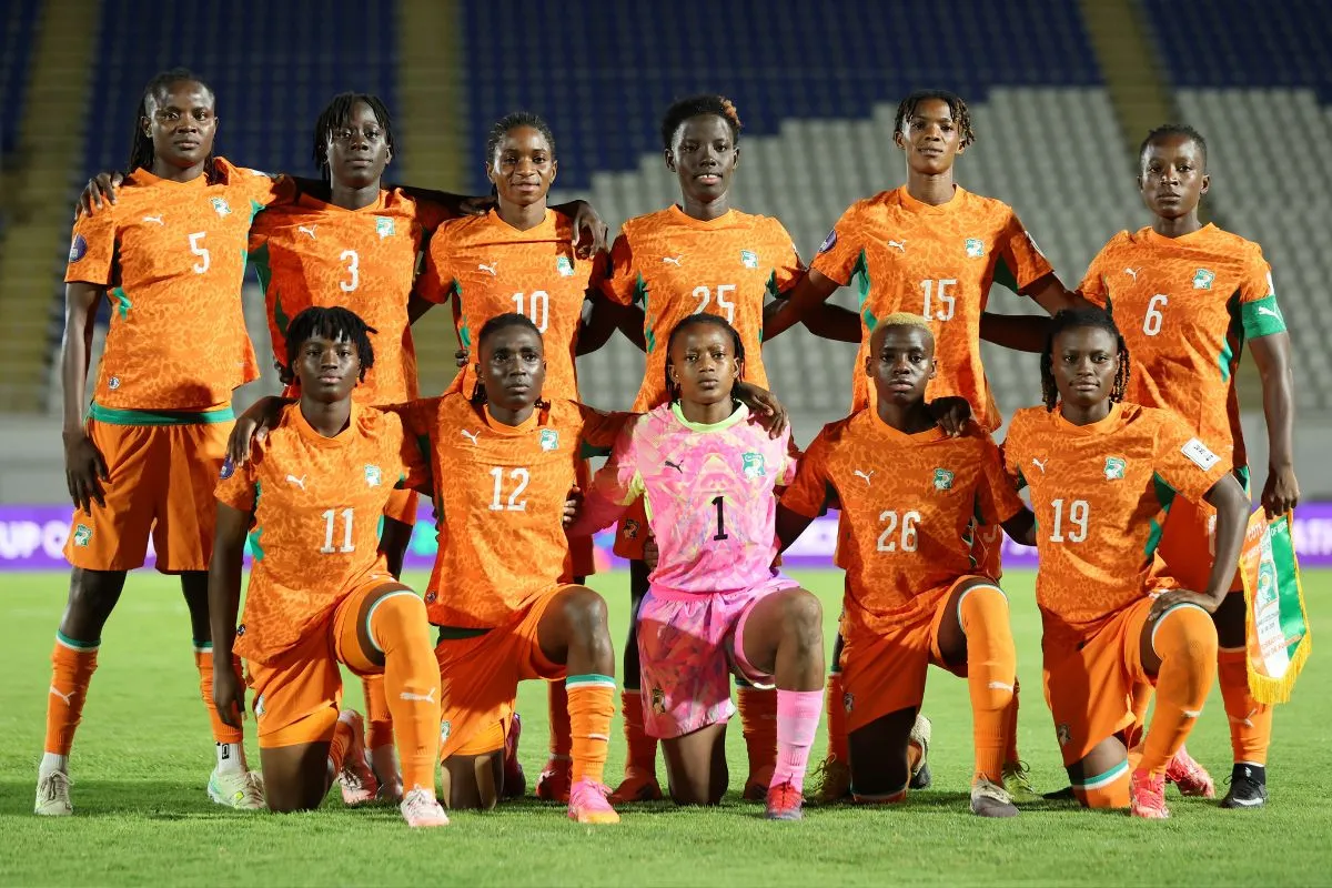 Eleven female soccer players in bright orange uniforms pose for a team photo on the field, with the goalkeeper in pink centered in front.
