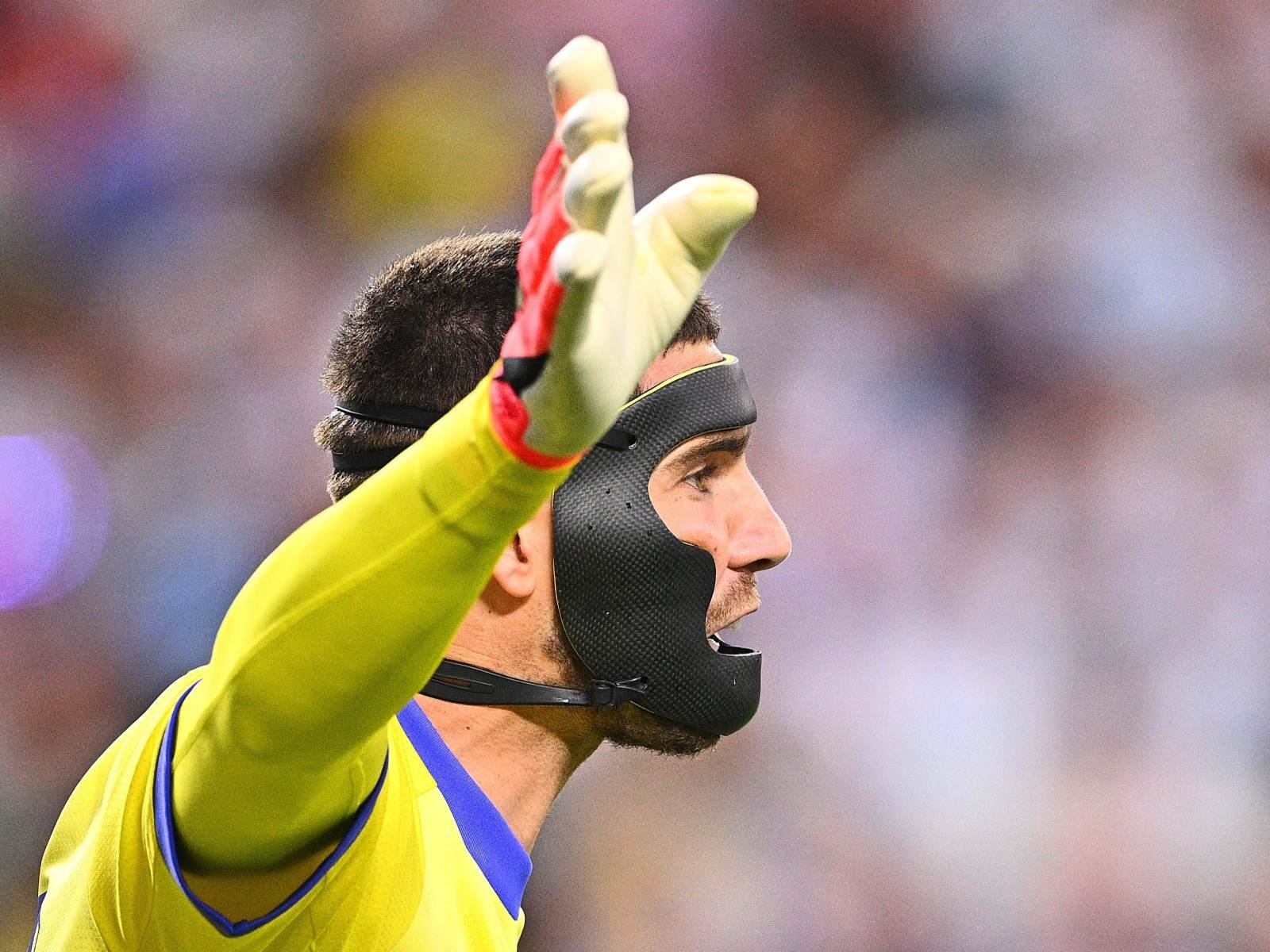 Soccer goalkeeper in a yellow jersey and black protective mask raises his gloved right hand.