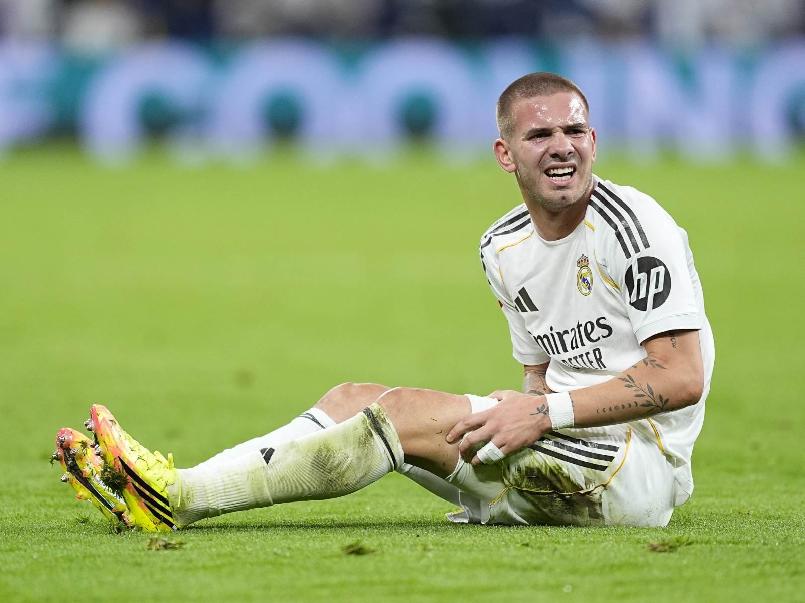 Soccer player in a Real Madrid white kit sitting on the grass, grimacing in pain after a challenge, yellow cleats visible.