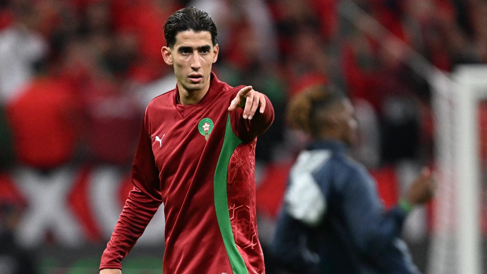 Soccer player in a red uniform with a green stripe points toward something on the field.