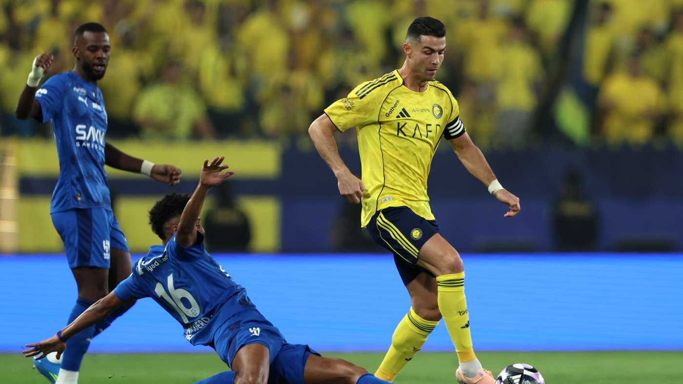 Soccer player in a yellow kit dribbles the ball past a sliding opponent in blue on a bright stadium field with a blurred yellow crowd in the background.