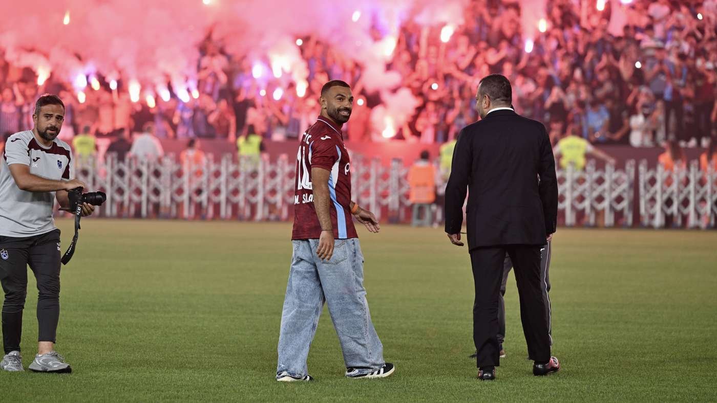 Soccer player in a maroon jersey and baggy jeans walks on the field as fans light red flares in the stands behind him.