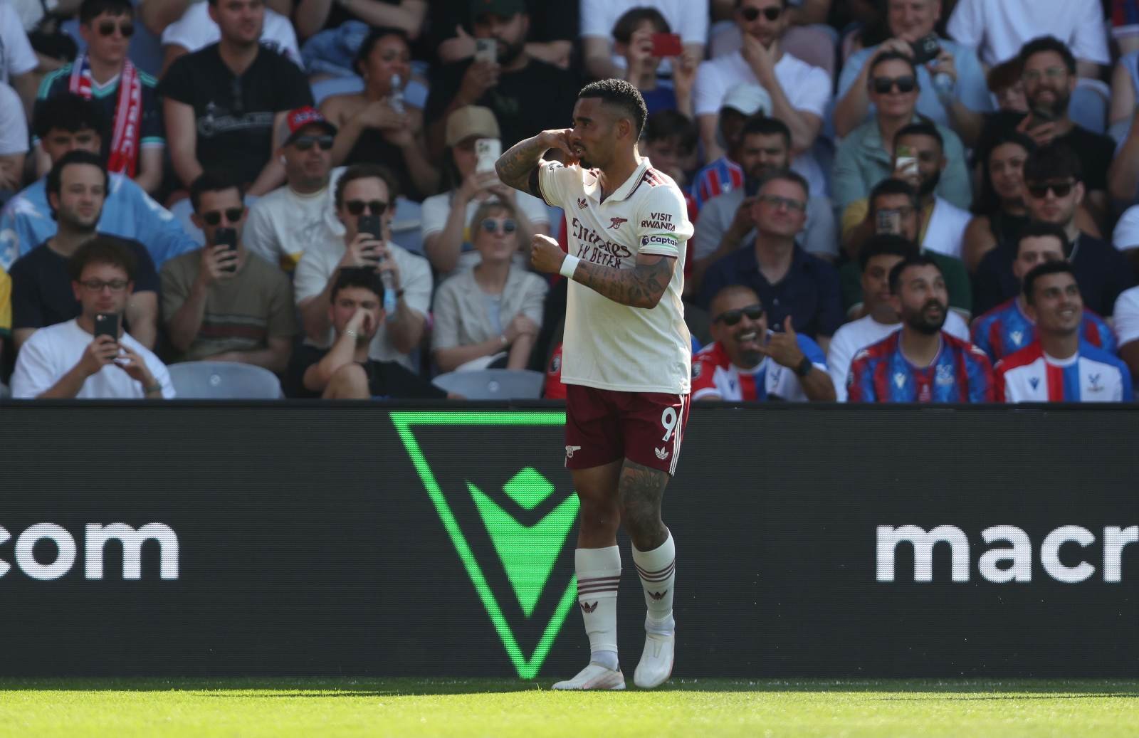 Soccer player in white jersey and maroon shorts, number 9, celebrating a goal on the pitch as fans film from the stands behind him.