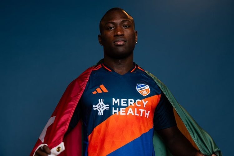Portrait of a Black male soccer player wearing a blue and orange Mercy Health FC Cincinnati jersey, draped with a flag over his shoulders.