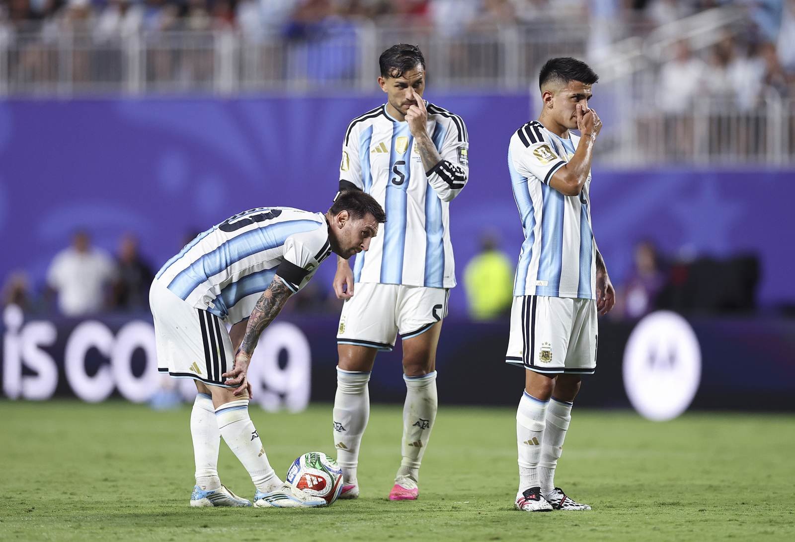Three Argentina players in light blue and white striped kits stand on a soccer field around a ball, one bending to reach it.
