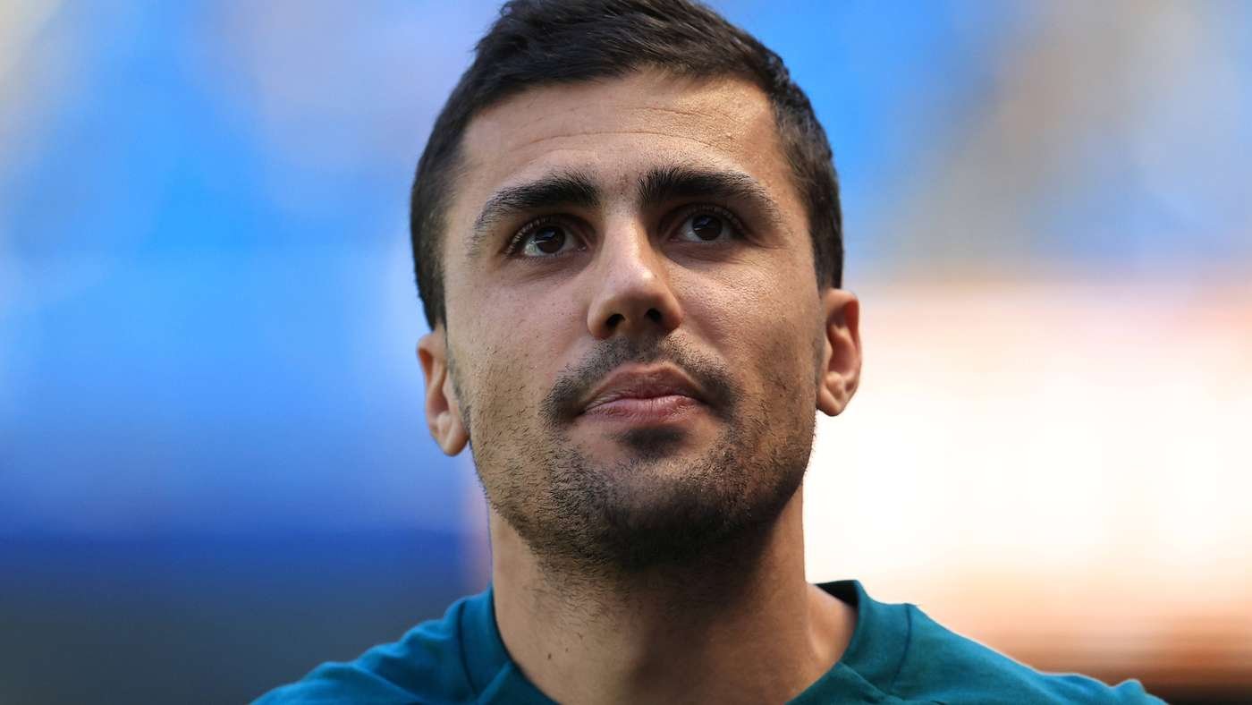 Close-up portrait of a man with short dark hair and stubble, looking upward against a blue blurred background.