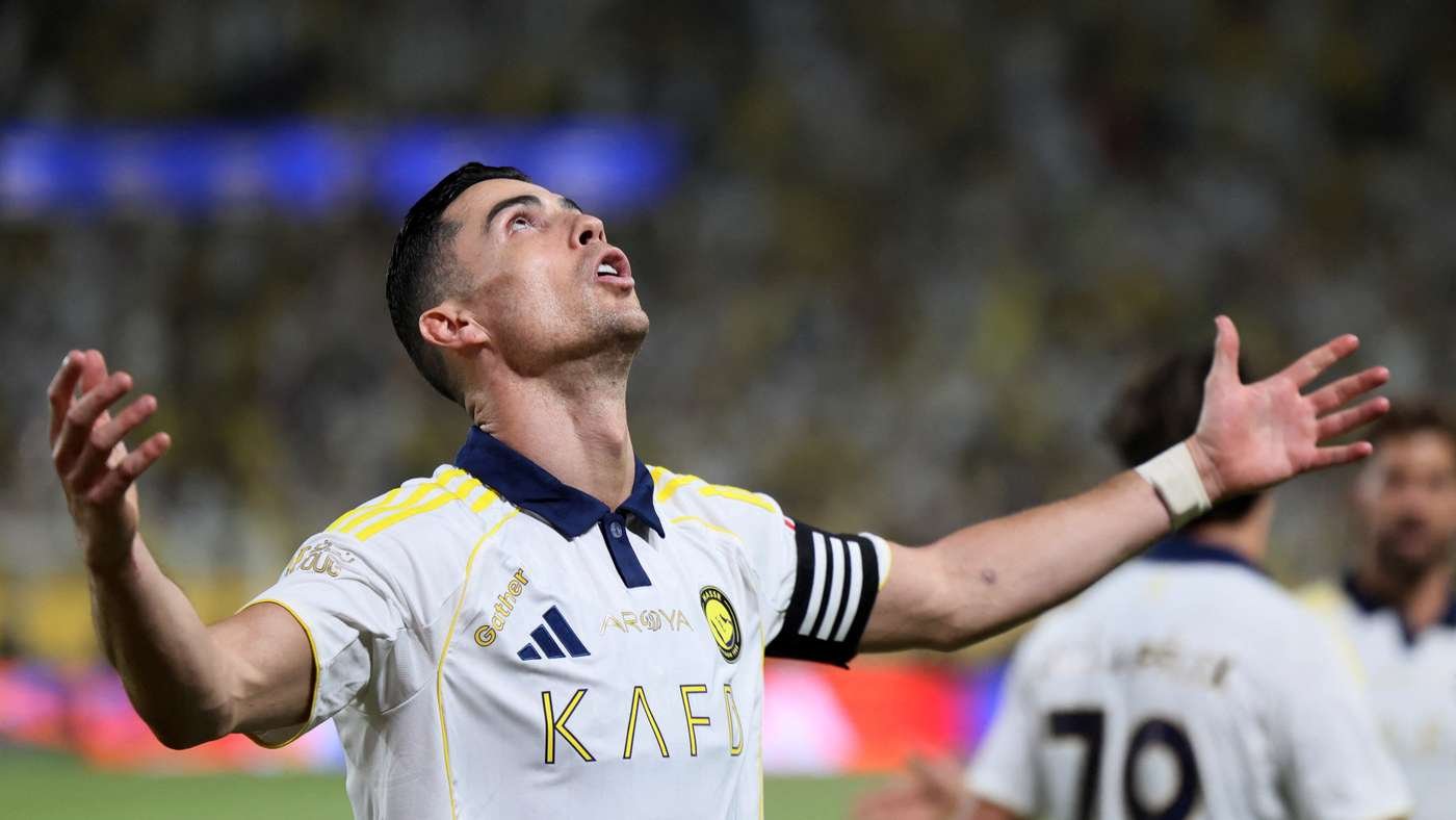 Soccer player in a white jersey with yellow accents and a captain's armband celebrates with arms raised, looking up.