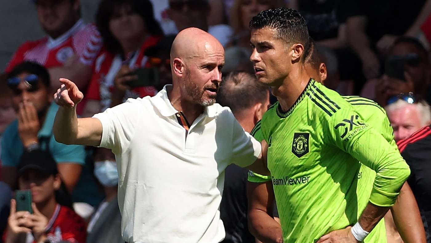 Football coach points and speaks to a Manchester United goalkeeper in a bright green kit during a match, with fans in the stands behind them.