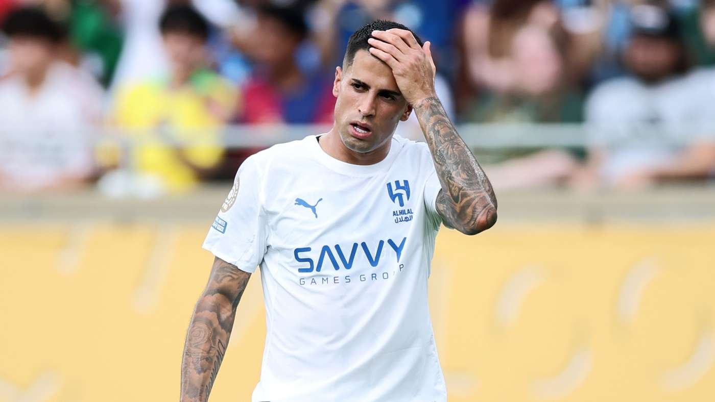 Soccer player in a white jersey touching his head, crowd blurred in the background, appears frustrated or tired.