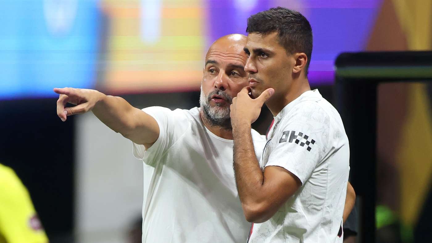 Coach with a shaved head and gray beard points off-frame while explaining strategy to a young player during a game.