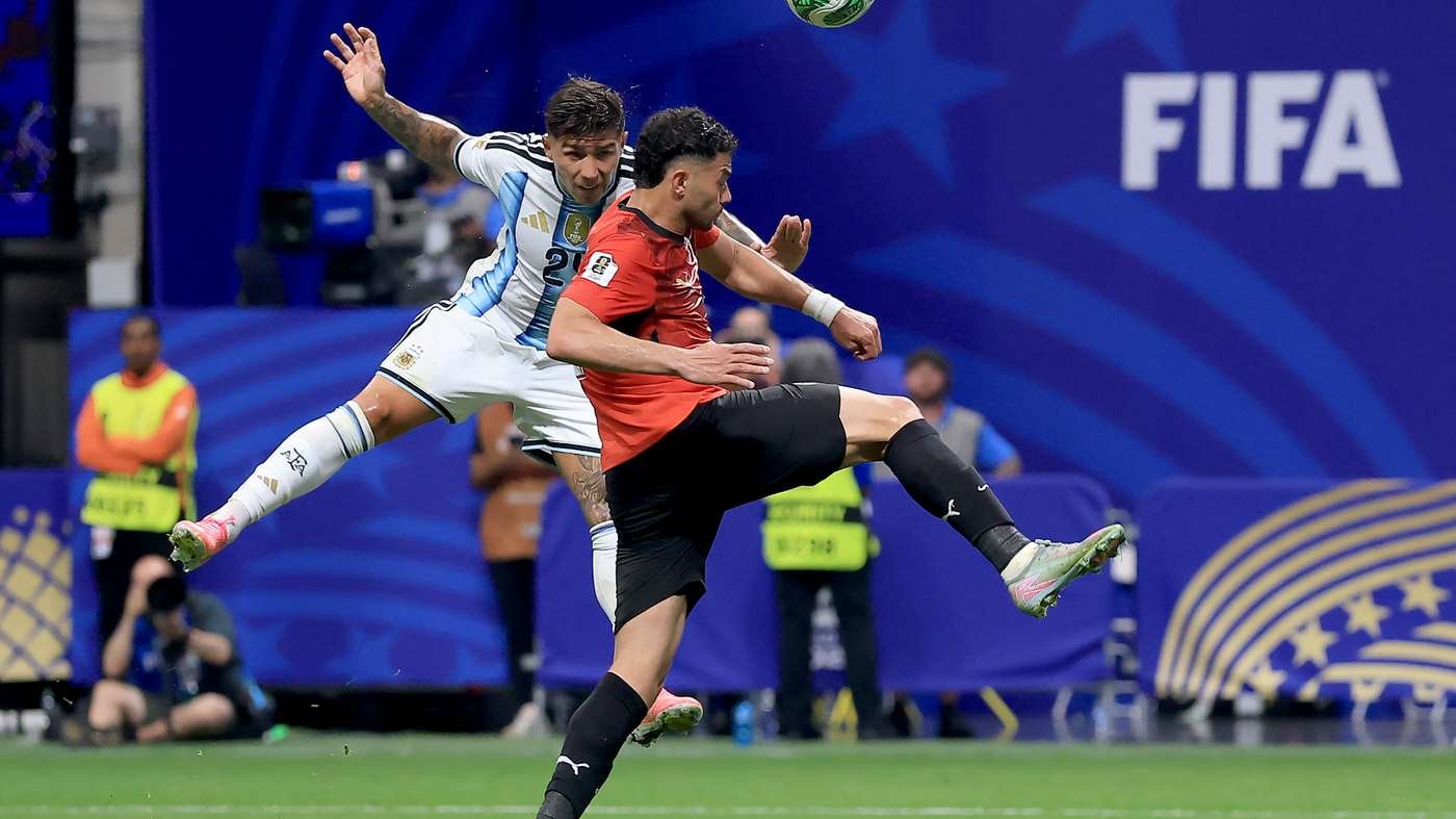 Two soccer players leap for the ball in a midair challenge; one in Argentina blue-white kit, the other in red, with a FIFA banner in the background.