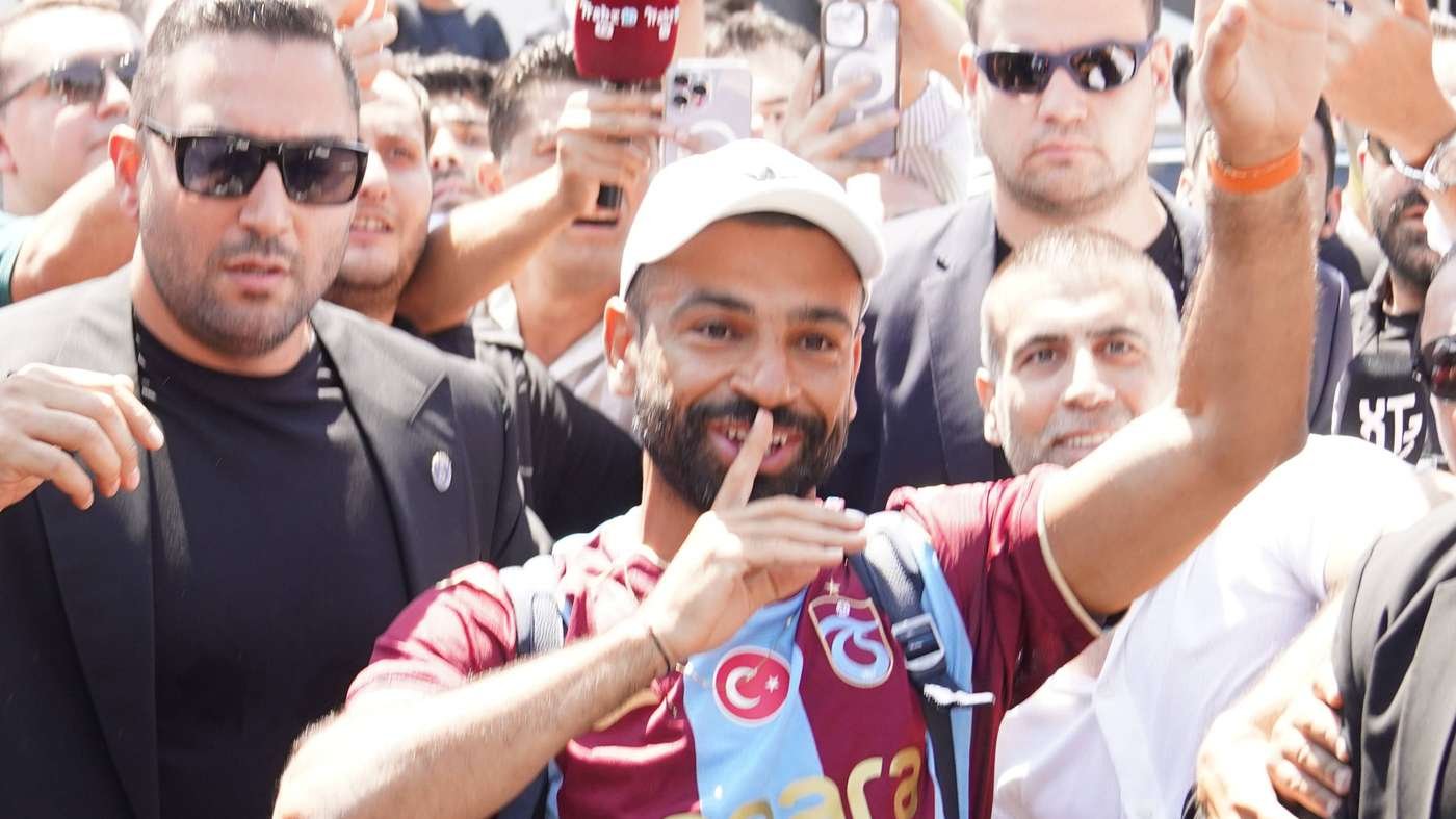 Man wearing a white cap and maroon-blue jersey smiles as fans crowd around taking photos and videos behind him.
