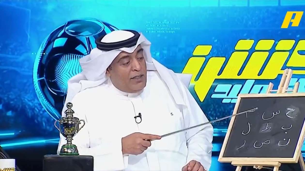 Man in white traditional dress points to a chalkboard displaying Arabic letters during a TV broadcast, with a trophy on the desk beside him.