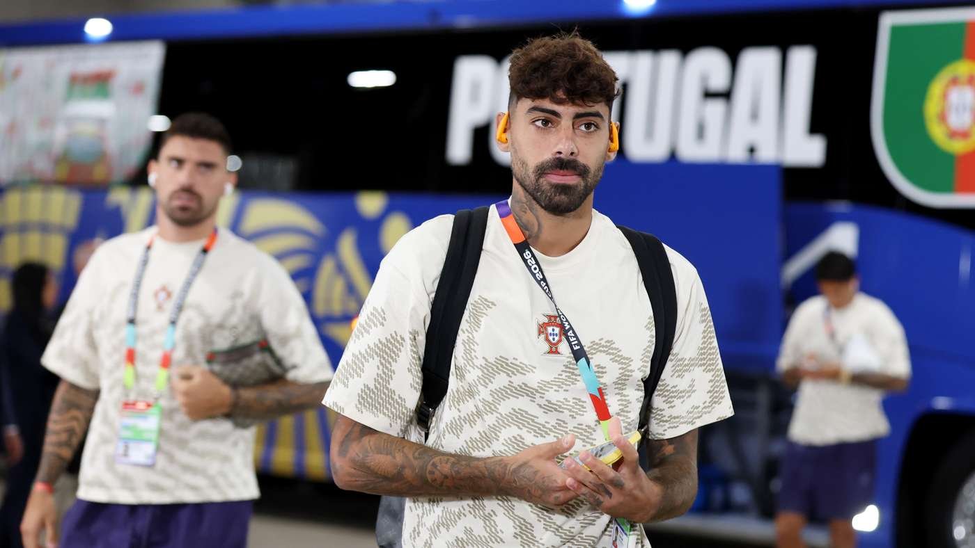 Man with tattoos and a beard wearing a Portugal crest shirt, a backpack, and orange ear protectors, standing in front of a blue bus with 'Portugal' text in the background.