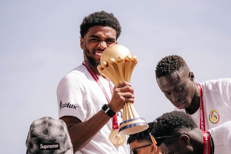 Young man on a podium holds a large gold trophy, smiling, with a medal around his neck amid teammates.