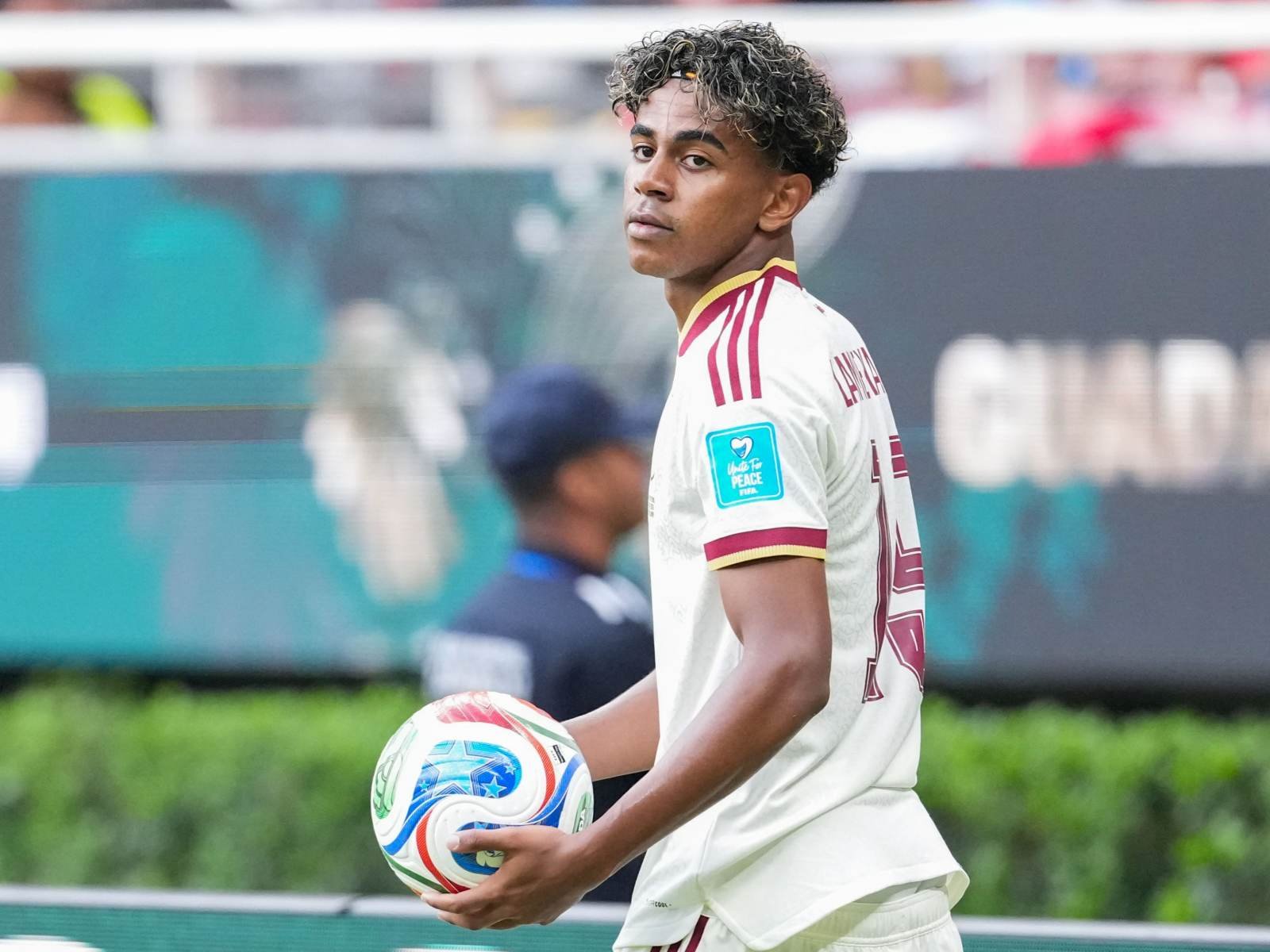 Soccer player in a white maroon-trimmed jersey holding a ball while looking toward the camera on a sideline.