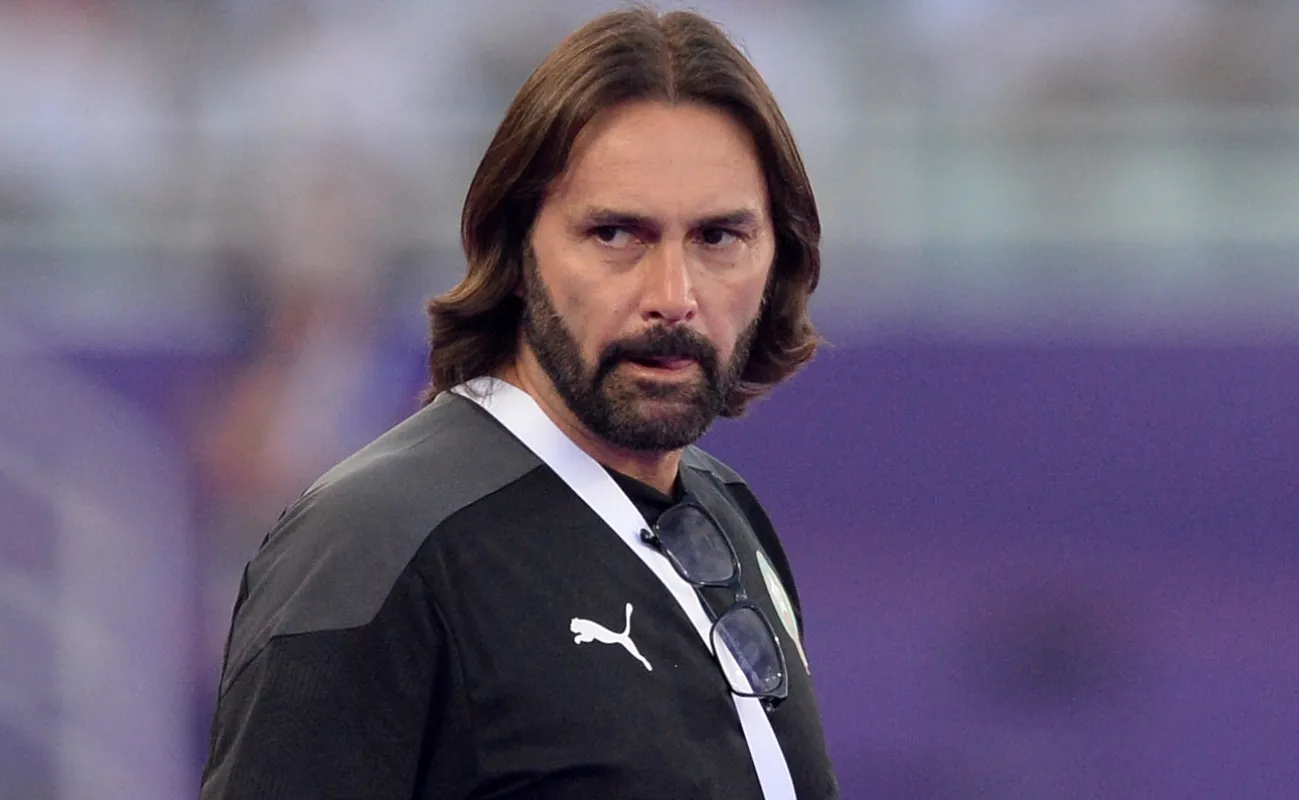 Man with shoulder-length brown hair and a beard, wearing a black Puma training shirt and glasses tucked at the chest, looking to the left in a stadium setting.