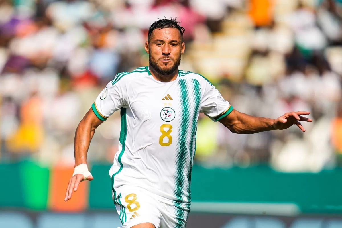 Soccer player in a white jersey with green accents and the number 8, sprinting on a sunny field with a blurred crowd.