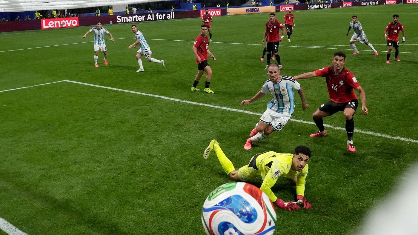 Goalkeeper in yellow on the grass reaching for the ball near the goal as an Argentina player and red-team opponents close in.