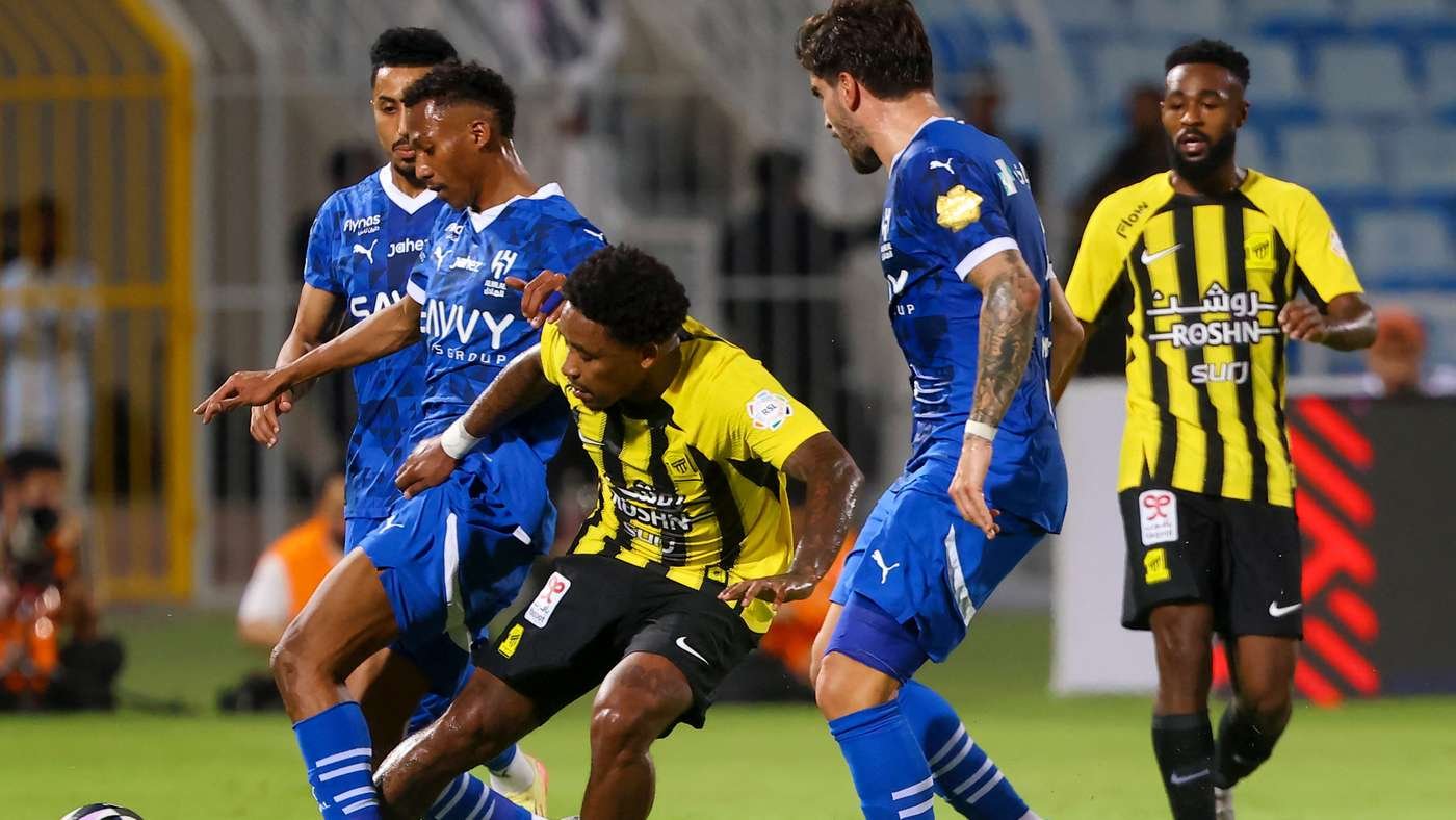 Soccer players in blue and yellow jerseys contest the ball during a match on a green field, two blue players close in on a yellow-and-black attacker.