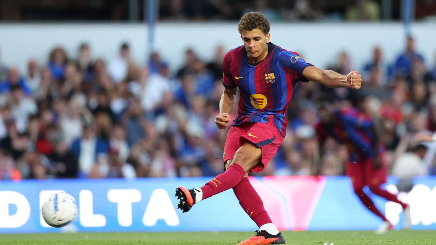 Barcelona footballer in action, kicking a ball during a match with a packed stadium in the background, wearing the blue and maroon kit.