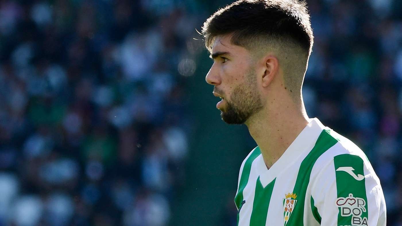 Soccer player in a green-and-white striped jersey, shown in profile with blurred stadium crowds behind him.