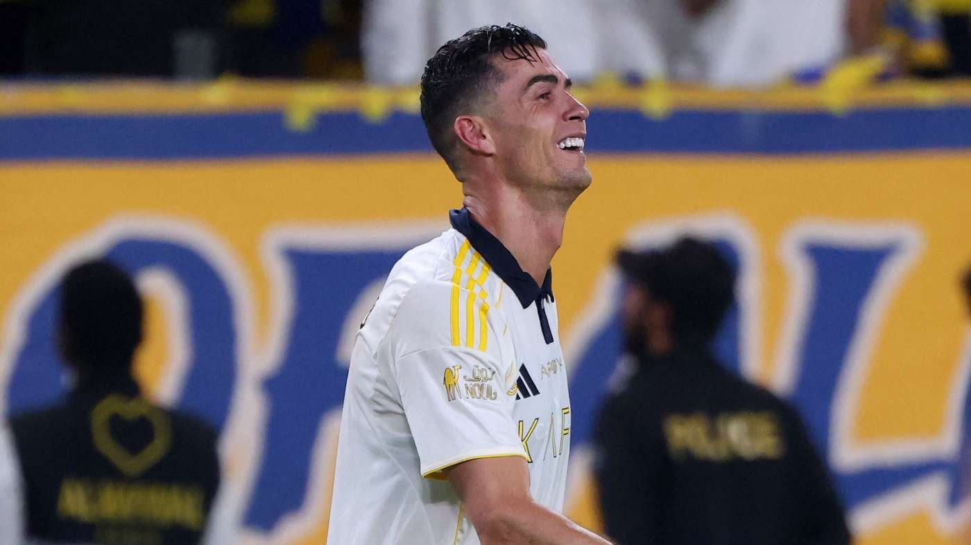 Soccer player in a white kit smiling on the field with yellow-blue stadium backdrop.