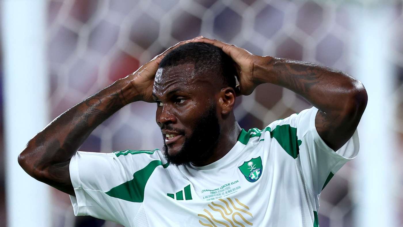 Soccer player in a white and green jersey holds his head in frustration on the field, sweat on his face.