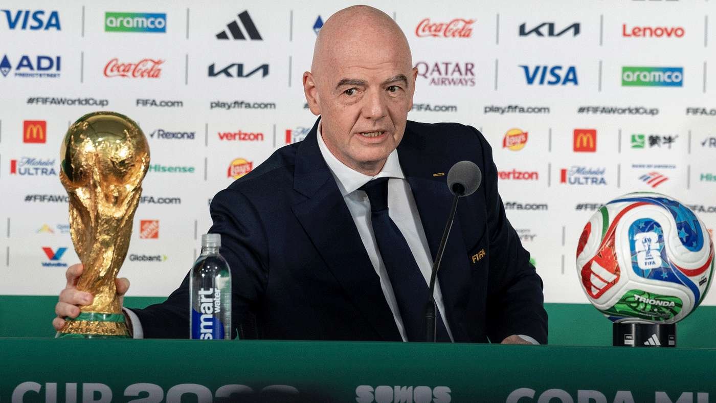 Bald man in a suit at a FIFA press conference, holding the World Cup trophy and a colorful soccer ball nearby.