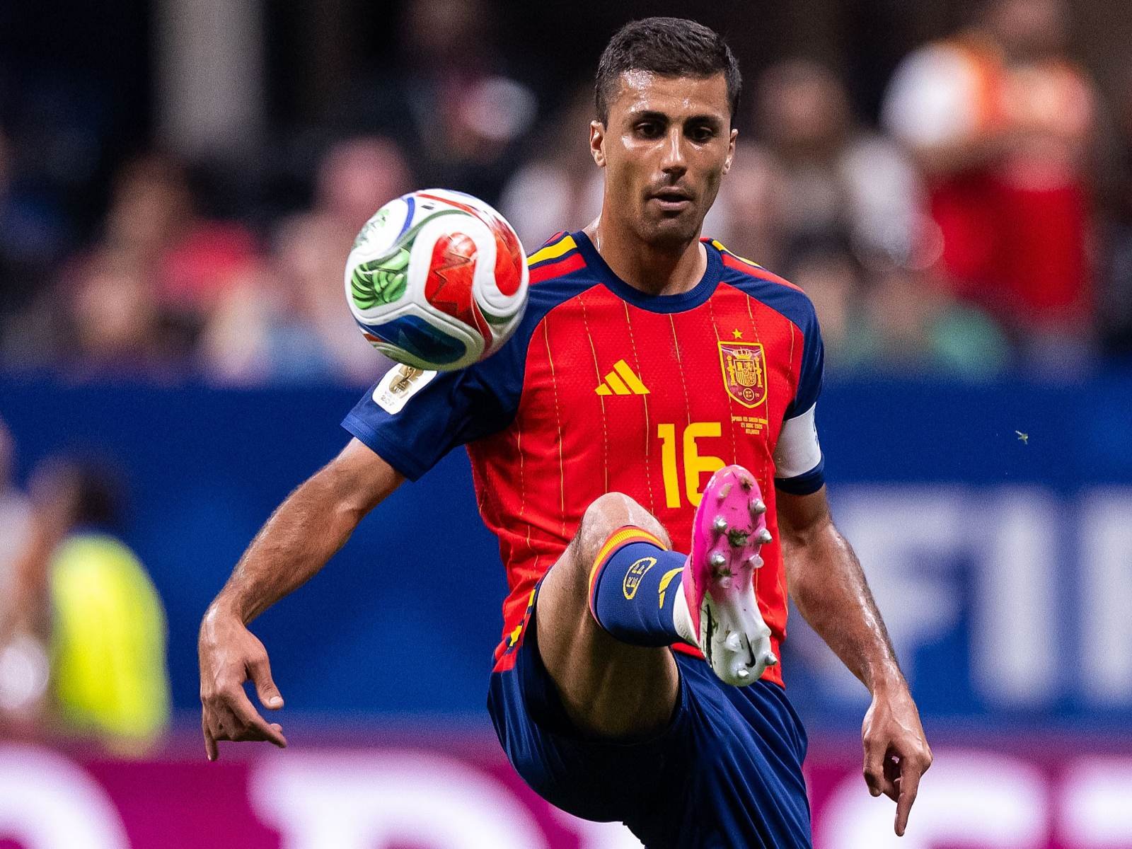 Soccer player in a red Spain jersey with number 16 controls a ball mid‑kick during a match, stadium crowd blurred in the background.
