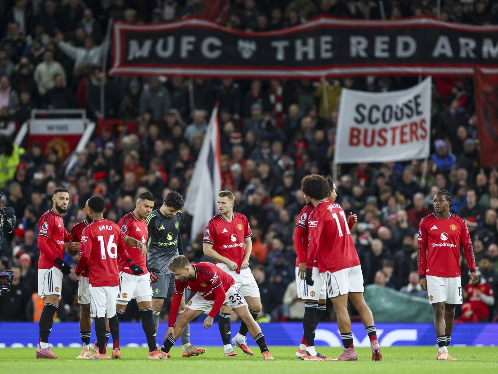 Manchester United players in red jerseys gather on the pitch during a game, with large banners in the stands reading 'MUFC' and 'SCOUSE BUSTERS' in the background. Manchester United players in red jerseys gather on the pitch during a game, with large banners in the stands reading 'MUFC' and 'SCOUSE BUSTERS' in the background.