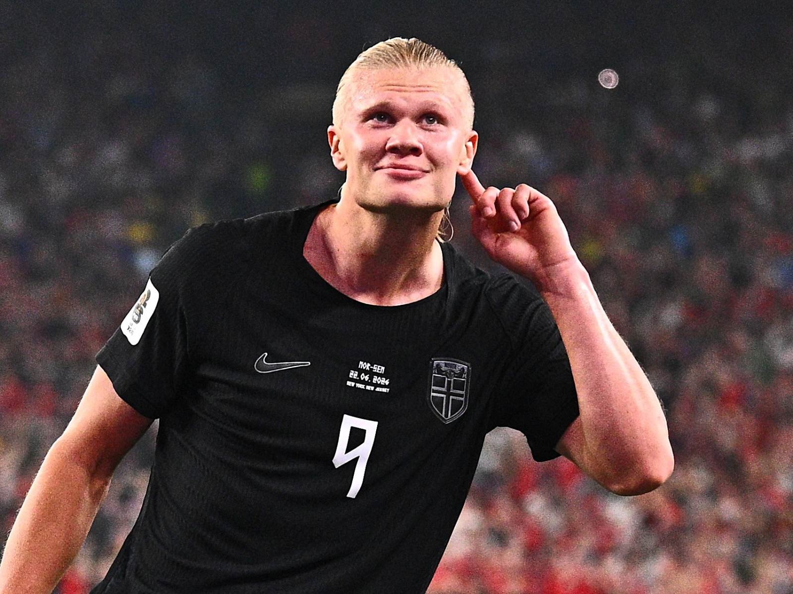 Soccer player in a black jersey with number 9 celebrating on the field, hand to ear as crowd cheers in the background.