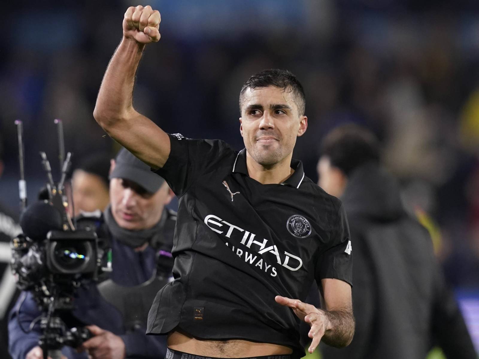 Soccer player in a black jersey raises a clenched fist in celebration on the pitch, with a camera crew in the background.