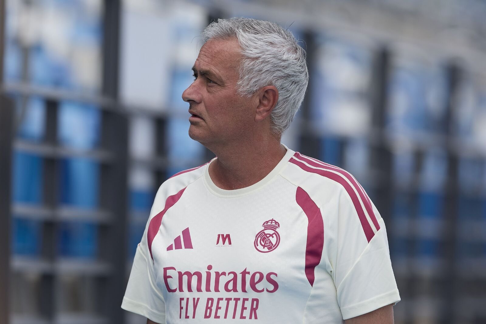 Man with gray hair in a Real Madrid jersey facing left outdoors with a blurred blue background.