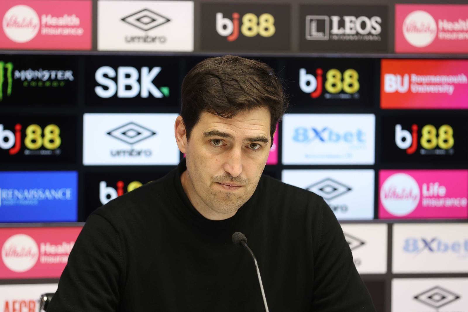 Man in a black shirt at a press conference, a microphone in front, sponsor logos on a backdrop behind him