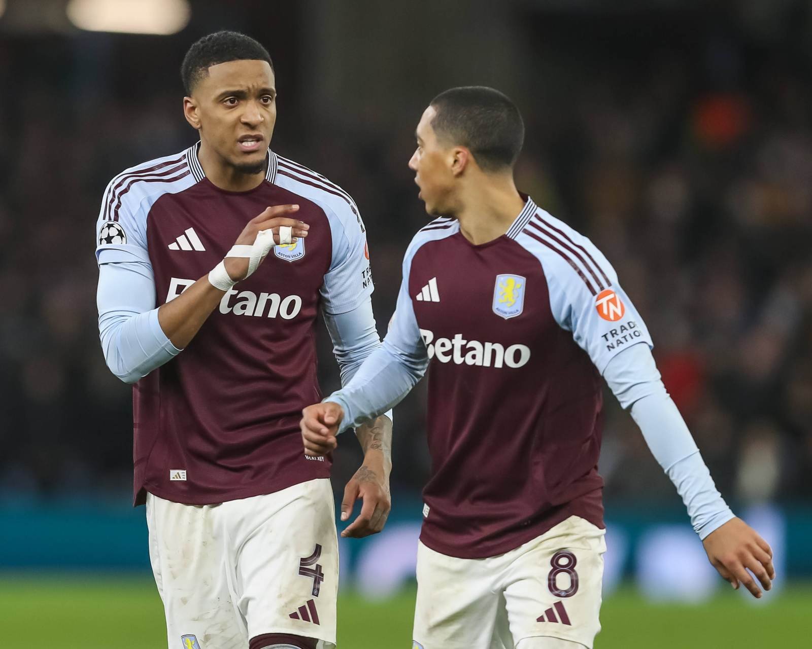 Two Aston Villa players in maroon and light-blue jerseys talk as they jog on the soccer pitch during a match.