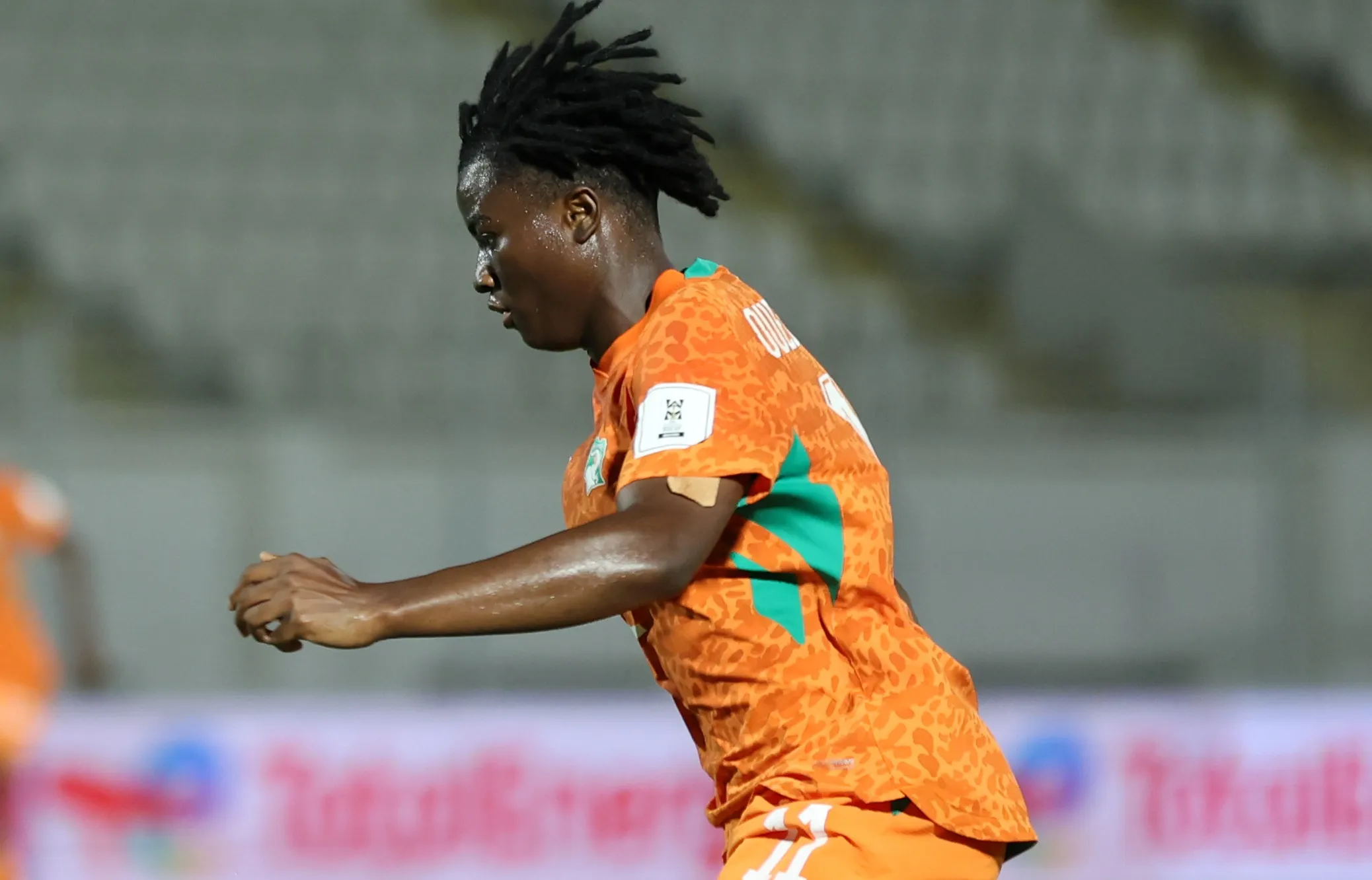 Soccer player in an orange kit running on the field during a match, side profile view with dreadlocks flying.