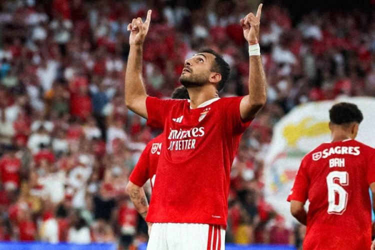 Soccer player in a red jersey celebrates with arms raised toward the sky on the pitch. Crowds cheer in the background. Option 2: "Red-clad footballer celebrates a goal with arms raised as fans celebrate behind him." Option 3: "Club footballer in red kit raises both arms after scoring, with teammates and cheering fans in the stands." Option 4: "Forward in red jersey pointing upward in celebration, during a match with a packed stadium."