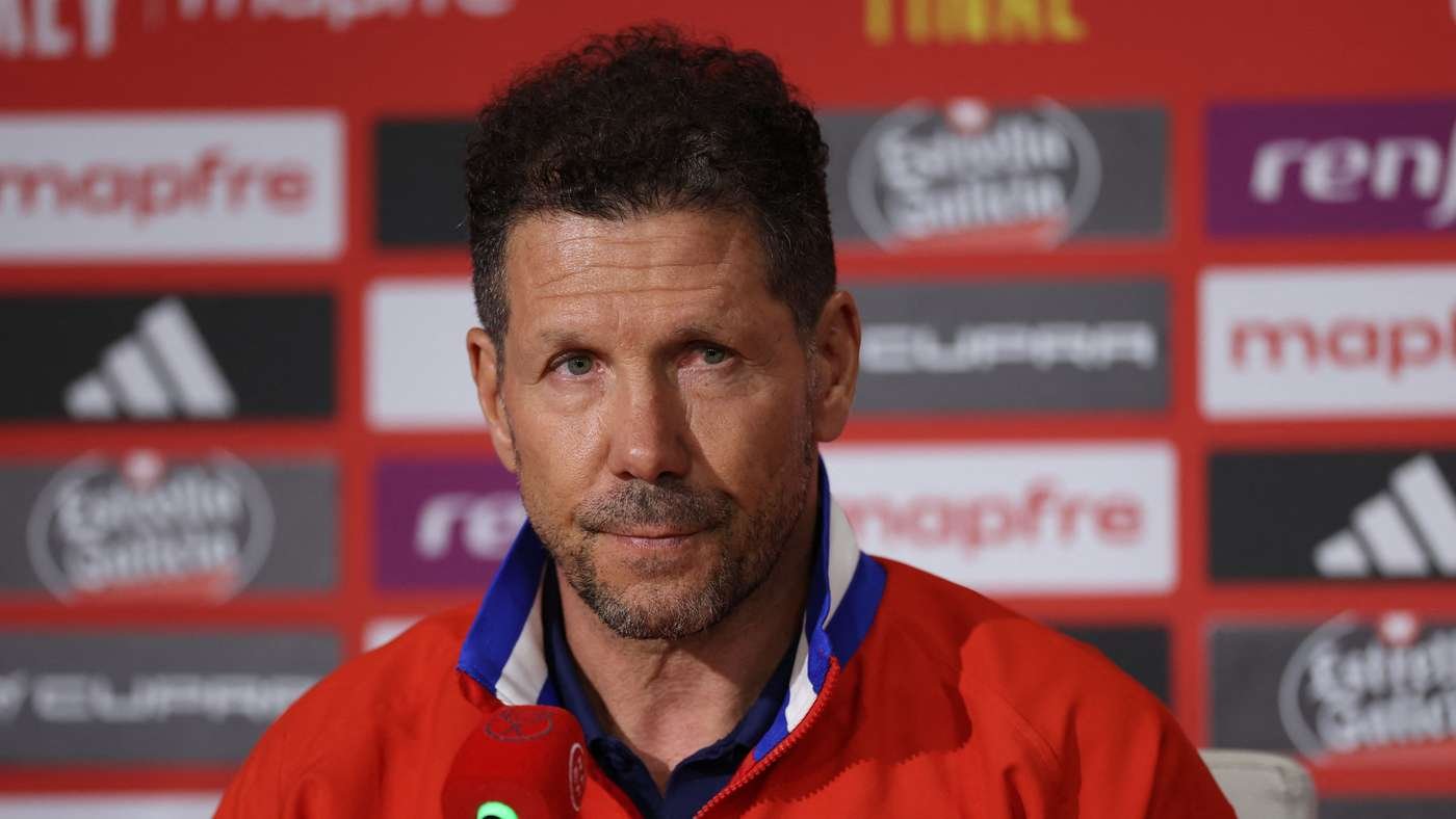 Man with short dark hair and stubble wearing a red jacket at a press conference backdrop filled with sponsor logos on a red wall.