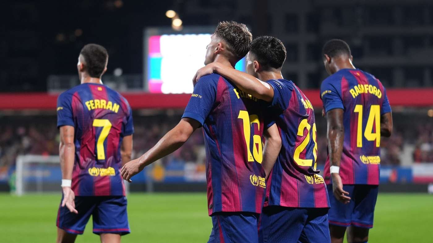 Soccer players in blue and maroon jerseys celebrate on the field, two with arms around each other in a night match.