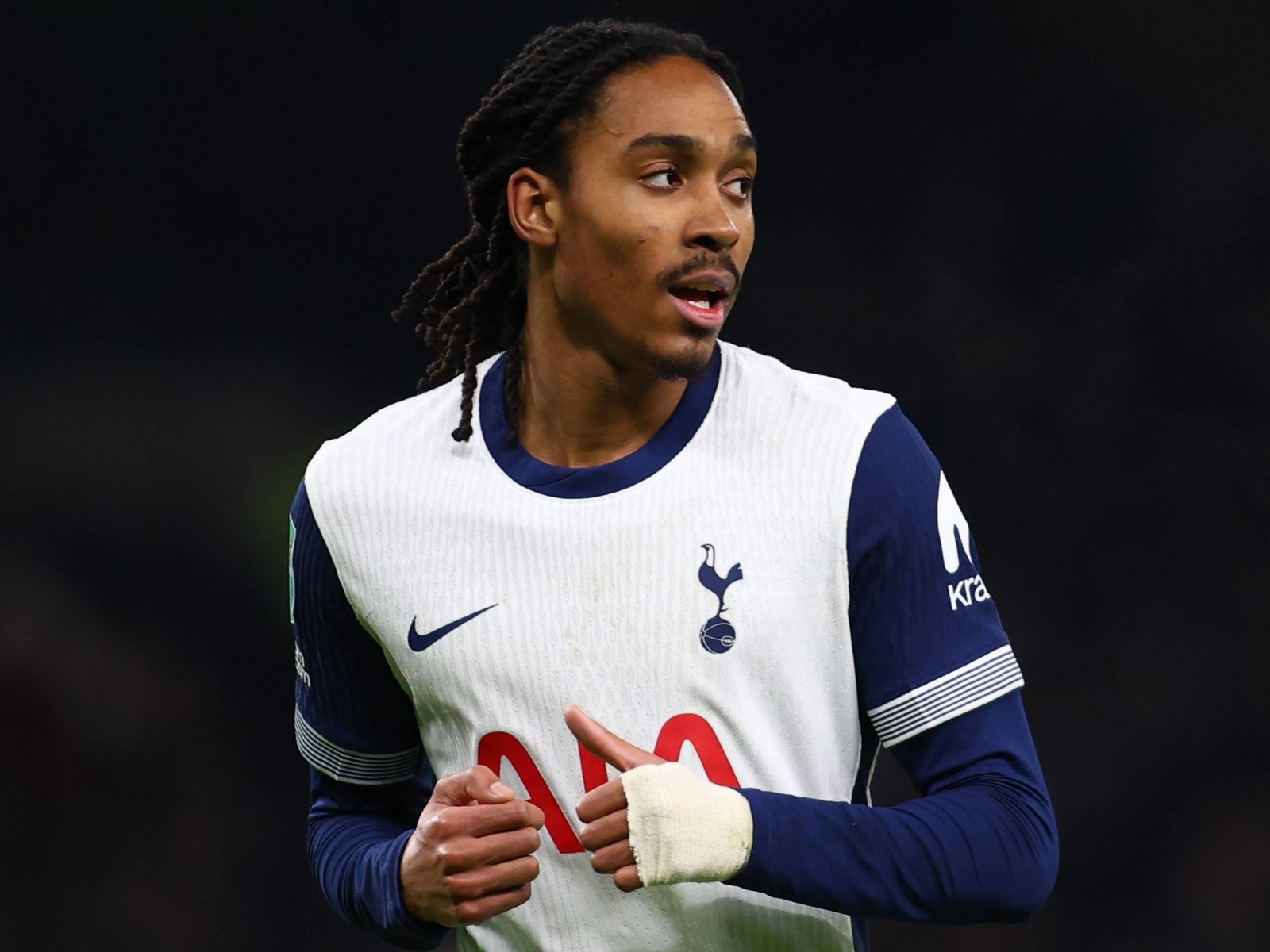 Soccer player with braided hair wearing a white training bib over blue jersey, holding a red card in front of a dark stadium background.
