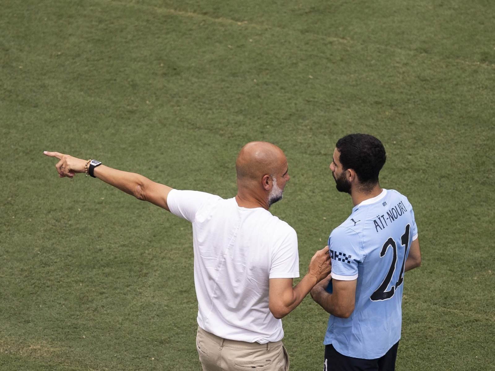 Coach in a white shirt points to the left while speaking to a footballer in a light blue jersey on a grass field behind them.