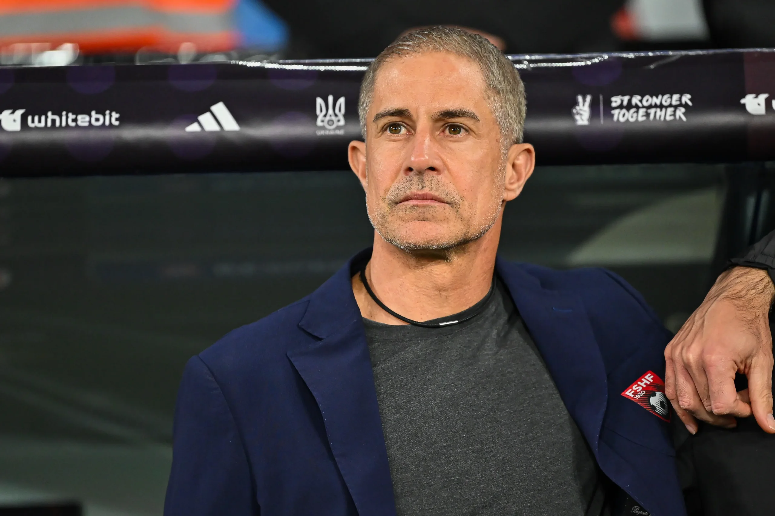 Man with short gray hair wearing a dark gray shirt and navy blazer, seated in a sports venue with sponsor banners behind him.