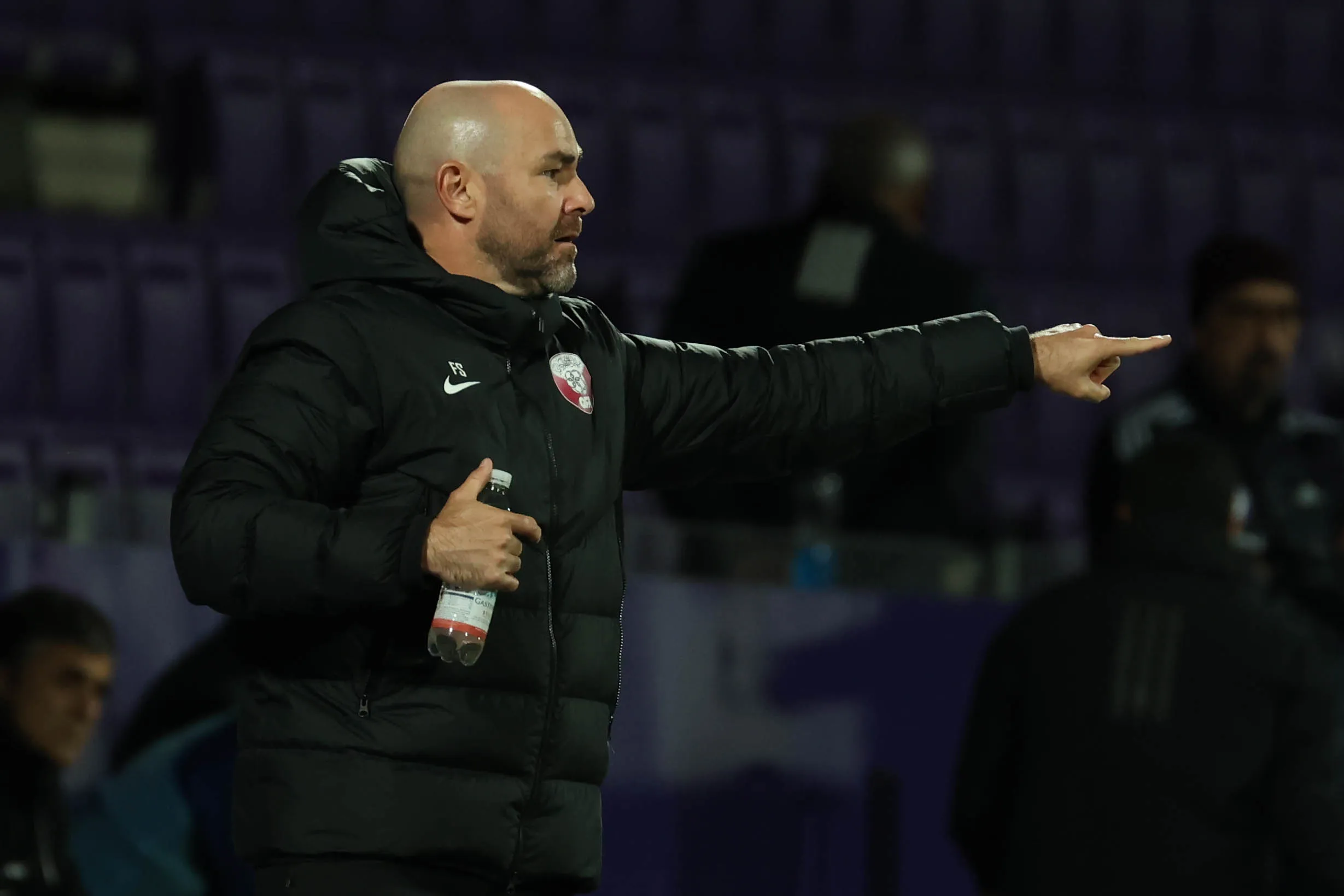 Coach-like man with a shaved head in a black puffer jacket points to the right while holding a water bottle outdoors at night stadium seating in the background