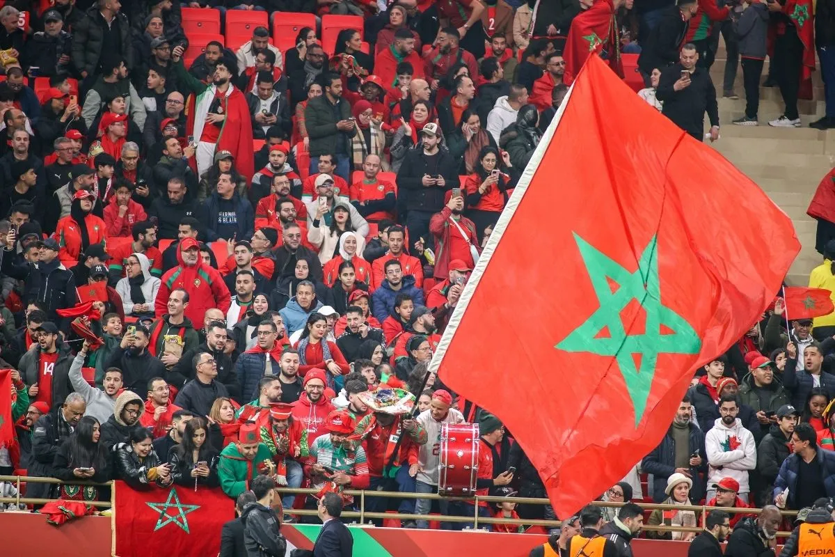 Crowd of football fans in red, with a large Moroccan flag waving in the foreground.